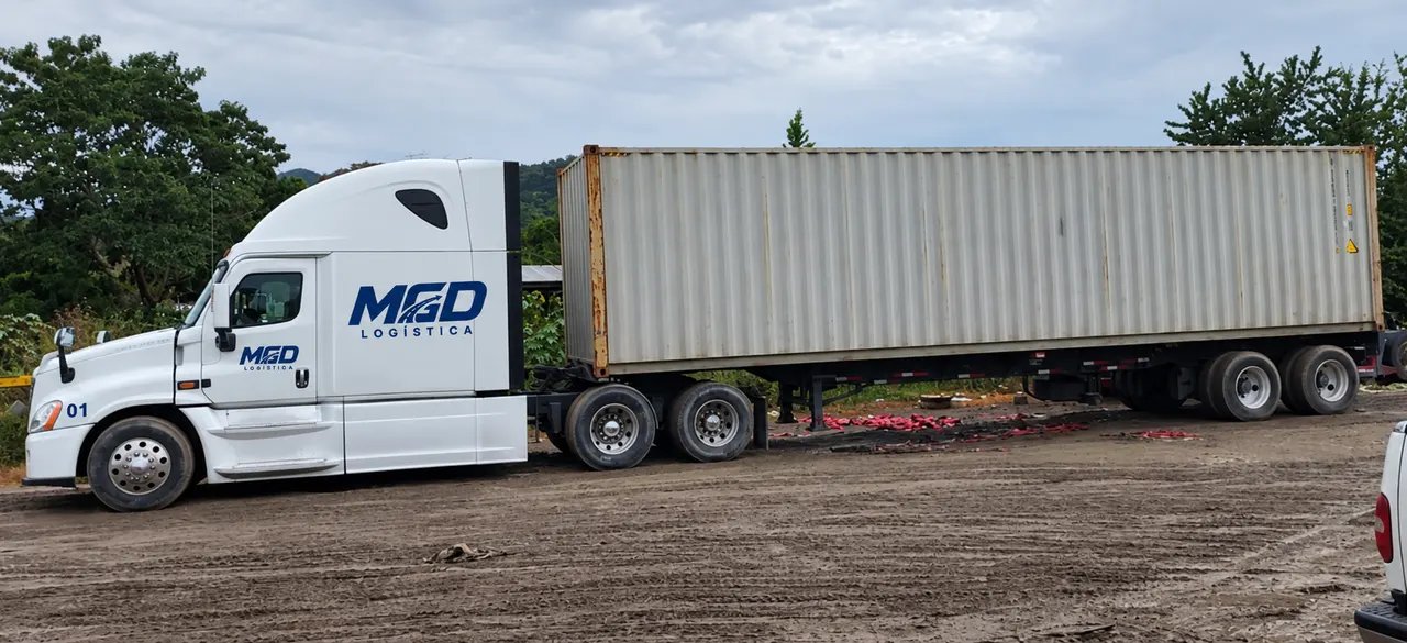Tractocamión blanco con el logo de MGD y un contenedor, en un patio de tierra