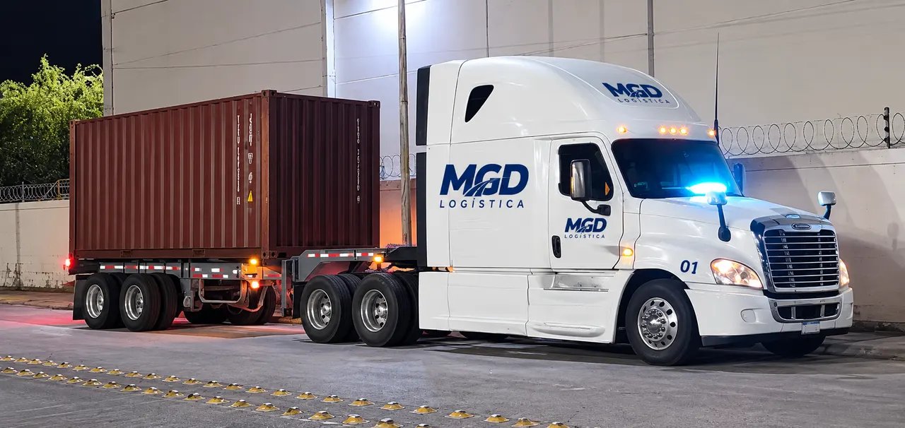 Tractocamión blanco con el logo de MGD y un contenedor en chasis, de noche junto a una nave iluminada