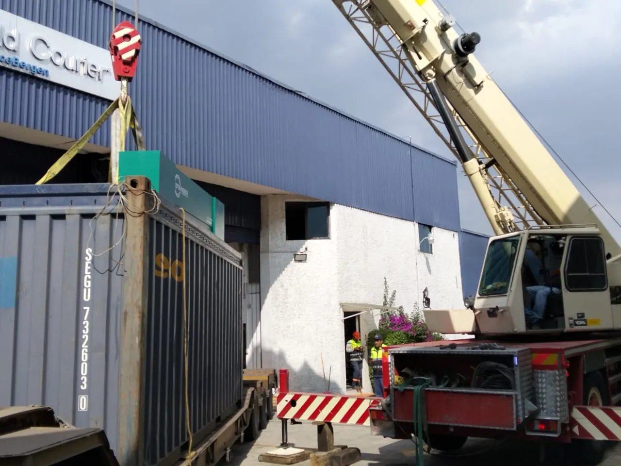 Grúa descargando con eslingas una pieza industrial de un contenedor open top, frente a una nave, con dos trabajadores con chaleco