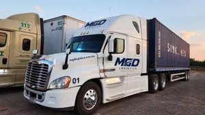 Tractocamión blanco con el logo de MGD y un contenedor de 40 pies, estacionado junto a otras unidades al atardecer